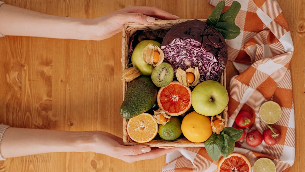 Vibrant basket of fresh fruits and vegetables with hands holding on a wooden table.